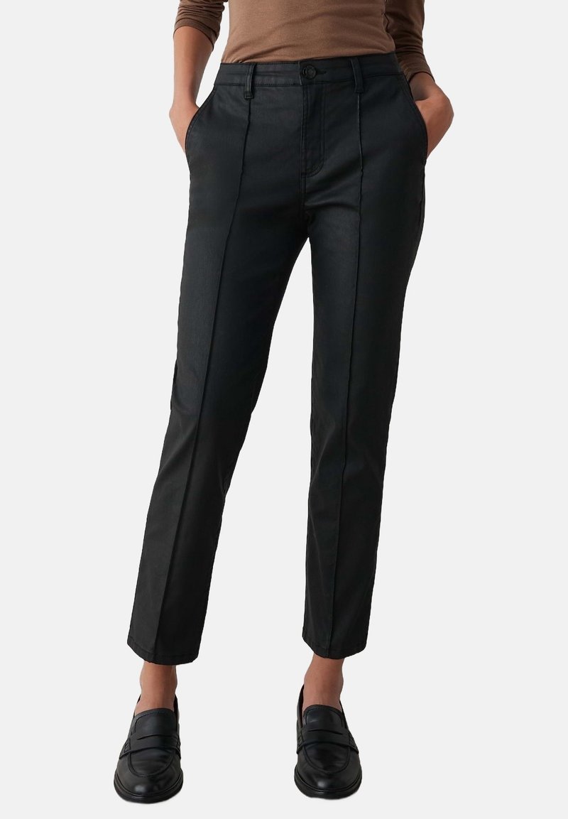Femme portant un pantalon slim noir avec des coutures devant, un haut marron à manches longues et des chaussures noires sans lacets, mains dans les poches.