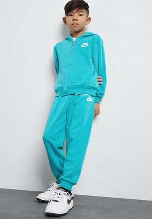 Sudadera Nike turquesa con cremallera delantera y logo, combinada con pantalones jogger a juego. Zapatillas blancas con acentos negros completan el conjunto.