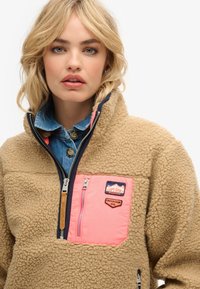 Tan fleecejakke med kvart lynlås, der har en lyserød brystlomme og patches. Kraven har et kontrasterende lag af blå denim nedenunder.