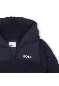 Veste bleu marine avec une capuche matelassée, texture côtelée et logo BOSS en blanc. Comprend une fermeture éclair et une doublure en polaire douce.