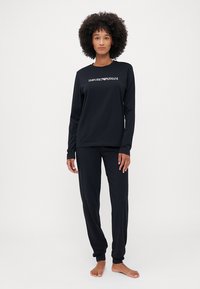 Melna garām piedurknēm kreklā un saskaņotās biksēs, kas izgatavotas no mīksta auduma. Krekls iekļauj baltu "Emporio Armani" logotipu. Šaura piegriezuma dizains, bez rakstiem.