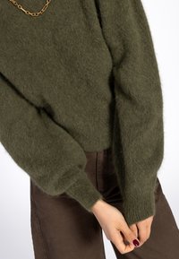 Olijfgroene pluche trui met lange, volumineuze mouwen en geribbelde manchetten, gecombineerd met donkerbruine broek. Bevat een eenvoudige gouden ketting.