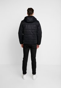 Svart vadderad jacka med huva, med en quiltad design. Bärs över svarta byxor och vita sneakers, vilket visar en avslappnad passform bakifrån.