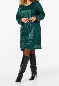 Smaragdgrön paljettklänning med långa ärmar, rund halsringning och hel passform. Kombinerad med svarta knähöga stiletto stövlar.