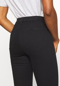 Jeans negros de cintura alta con una textura suave, dos bolsillos traseros y detalles de costura sutiles. La tela parece elástica y ajustada al cuerpo.