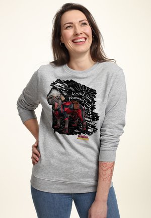 Femme souriante portant un sweat-shirt gris avec un chien cartoon en costume rouge et le texte « On dirait une dinde plumée » de Deadpool vs Wolverine.