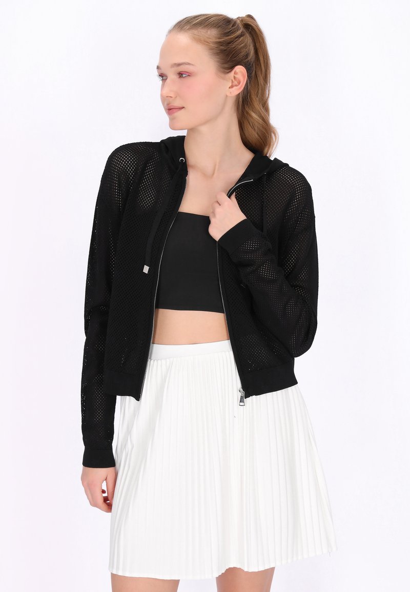 Jonge vrouw met een paardenstaart, gekleed in een zwarte mesh hoodie met ritssluiting over een zwart crop top en een witte geplooide rok, kijkt opzij.