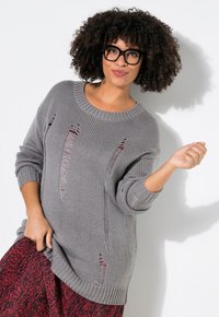 Grauer Strickpullover mit geripptem Ausschnitt und oversized Schnitt, der mehrere abgenutzte Stellen aufweist. Kombiniert mit einem rot gemusterten Rock.