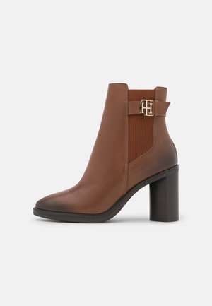 Bota de cuero marrón hasta el tobillo con punta puntiaguda, paneles laterales elásticos y hebilla de cinturón con detalle en dorado. Tacón grueso y texturizado. Acabado liso.