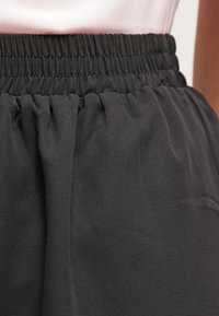 Musta joustava vyötäröinen hame, jossa kerätty pinta, sileä kangas ja hienovarainen kiilto. Siinä on leveä vyötärönauha ja virtaava muotoilu.