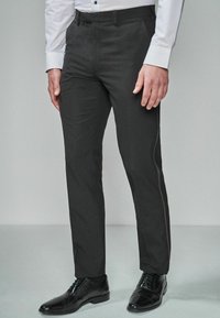 Pantalon noir ajusté, fabriqué dans un tissu lisse, avec un léger éclat et un design épuré, associé à des chaussures noires.