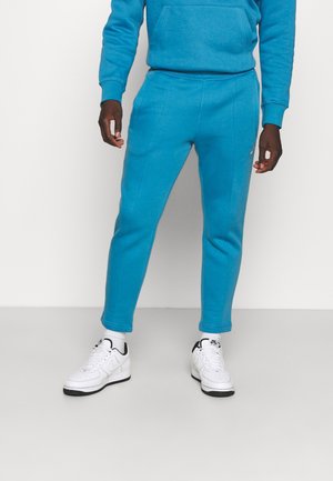 Hombre vestido con pantalones joggers azul brillante, sudadera con capucha a juego, calcetines blancos y zapatillas Nike blancas, de pie frente a un fondo liso.