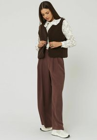 Gilet en polaire marron porté sur un blouse blanche à motifs floraux, associé à un pantalon maroon ample et des baskets blanches. Le gilet présente des détails de boutons.