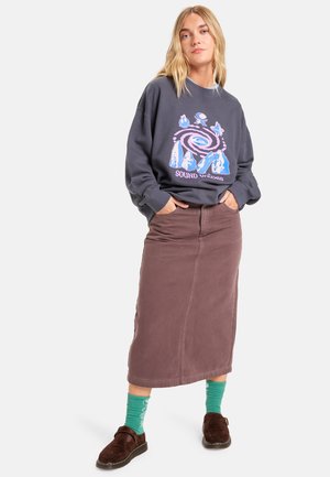 Sweatshirt gris avec un graphique coloré, jupe midi marron, chaussettes vertes à motifs et sabots marron foncé. Tenue décontractée avec des textures variées.