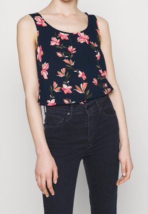 Frau trägt ein ärmelloses, marineblaues Oberteil mit pink-orangefarbenem Blumenmuster, das in eine hoch taillierte schwarze Jeans gesteckt ist, vor einem einfarbigen Hintergrund.