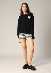 Pull en tricot noir avec un patch en forme de cœur blanc, associé à un short à carreaux noir et blanc. Le modèle porte des chaussures noires au style épais.