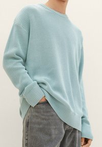 Osoba oblečená v světle modrém ribbovaném svetr s dlouhými rukávy, mírně zasunutém do vysokých šedých džín s vybledlým vzhledem, na neutrálním pozadí.