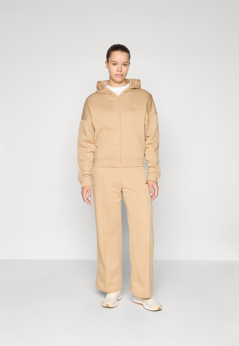 Beige zip-up college sweatshirt with kangaroo pockets and matching wide-leg pants. Made of soft material, it features a subtle logo on the chest and side.