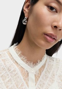 Orecchini d'argento con un design sferico, caratterizzati da una superficie liscia e lucida e una finitura opaca a contrasto. Indossati con una blusa in pizzo.