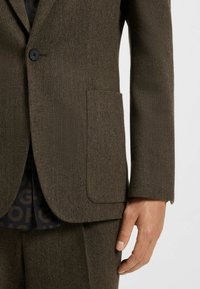 Brauner Fischgrat-Blazer mit einem einzelnen Knopf, aufgesetzten Revers und einer Brusttasche, kombiniert mit einem gemusterten dunklen Hemd darunter.