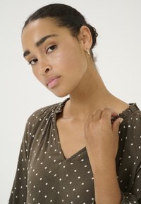 Blouse brune à pois avec un décolleté en V et un col froncé. Présente des manches amples et un tissu texturé ; modèle portant des boucles d'oreilles en or.