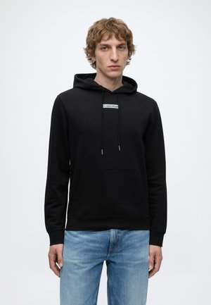 Young man wearing a black Calvin Klein hoodie with front pocket and light blue jeans, standing against a plain white background.