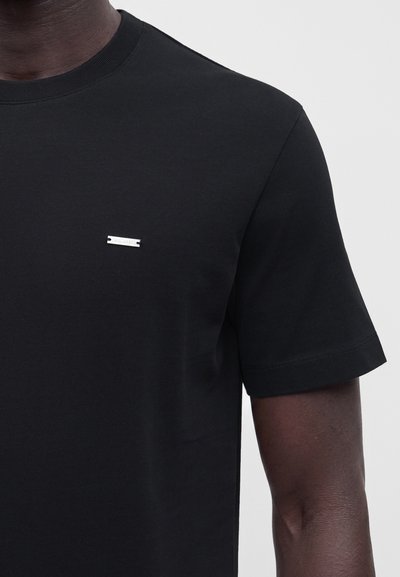 Homme portant un t-shirt noir uni à manches courtes avec une petite plaque de logo argentée horizontale sur le côté gauche de la poitrine, sur un fond clair.