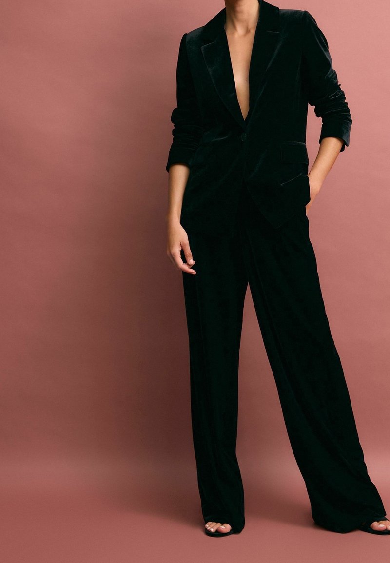 Personne vêtue d'un tailleur pantalon en velours noir avec un blazer à col en V profond et un pantalon à jambes larges, debout devant un fond rose atténué.