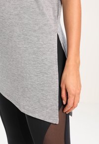 Main d’une femme reposant à côté de leggings noirs avec panneaux en mesh, portant un haut tunique gris clair avec une fente latérale.