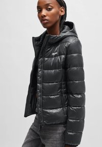 Schwarze Pufferjacke mit Kapuze, glänzender Oberfläche, gesteppter Optik, Frontreißverschluss und Seitentaschen. Weißes "Hugo"-Logo auf der Brust.
