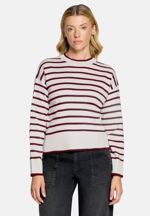 Kvinde med blondt hår iført hvid sweater med vandrette mørkerøde striber og højtaljede sorte jeans mod ensfarvet baggrund.