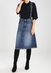 Person som bär en svart lös t-shirt, en blå denim hängselskör med franskant och svarta knähöga stövlar, som står mot en vit bakgrund.