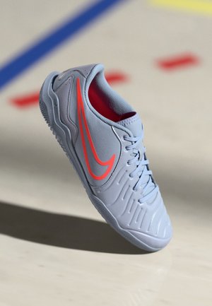 Zapatilla deportiva gris claro con logo swoosh naranja brillante y forro interior rojo sobre suelo pálido con líneas coloreadas.