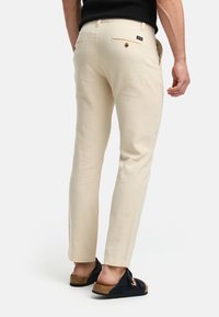 Beige linen pants with a slim fit, featuring a back pocket and logo label. Worn with dark slip-on shoes, showcasing a simple design.