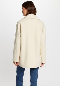 Cappotto crema in misto lana con ampio colletto, veste morbido e spalle scese. Caratterizzato da una texture liscia e un design di lunghezza media.