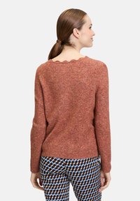 Rosenfarbener, gestrickter Pullover mit gewelltem Ausschnitt, langen Ärmeln und lockerer Passform. Kombiniert mit gemusterten Hosen, die geometrische Formen zeigen.