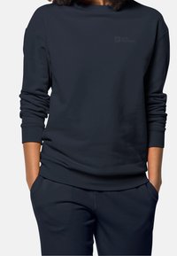 Löysä istuva tummansininen collegepaita pehmeästä kankaasta, leveällä kaula-aukolla ja käärityillä hihoilla. Rintakehässä hienovarainen logotyyppitys, yhdistettynä samaan sävyyn oleviin housuihin.