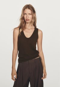 Frau mit gewelltem Haar, die ein ärmelloses schwarzes Top und eine hoch taillierte, lockere schwarze Hose vor einem schlichten hellen Hintergrund trägt.