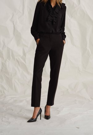 Blouse en soie noire à col volanté, associée à un pantalon noir ajusté et des escarpins noirs pointus à talons aiguilles, sur un fond blanc froissé.