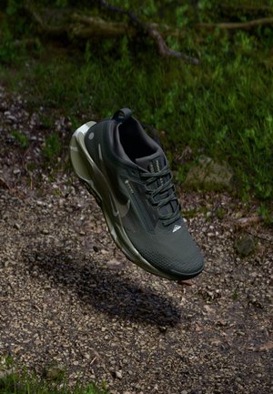 Scarpa sportiva nera e grigia con suola texturizzata che fluttua sopra un sentiero di ghiaia circondato da erba verde e piccoli sassi.