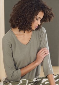 Olivgrünes V-Ausschnitt-Shirt mit Dreiviertelärmeln, das weißen "FRENCH CONNECTION"-Text auf dem Ärmel und eine weiche Stofftextur bietet.