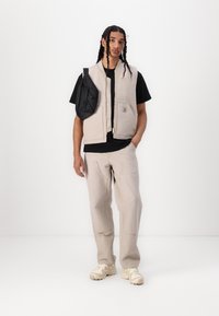 Beige polstret vest med glidelås foran og brystlomme, kombinert med en svart t-skjorte og lysebeige bukser. Tilbehøret inkluderer en svart bag.