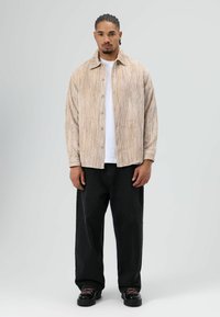 Overhemd van lichtbeige ribfluweel met knopen, gecombineerd met losse zwarte broeken. De outfit bestaat uit een wit T-shirt eronder en zwarte sneakers.