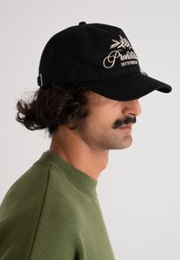 Svart caps med brodert tekst og bladdetalj. Har en buet skygge, solidt stoff og justerbar stropp bak. Grønn genser synlig.