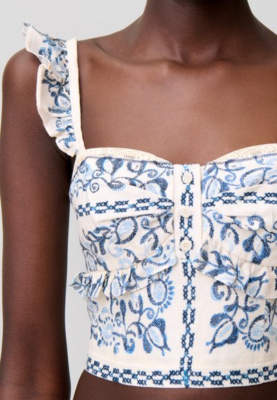 Crop top bianco senza maniche con ricamo floreale blu, apertura frontale con bottoni, spalline arricciate e dettaglio a volant sotto il seno su persona dalla pelle scura.