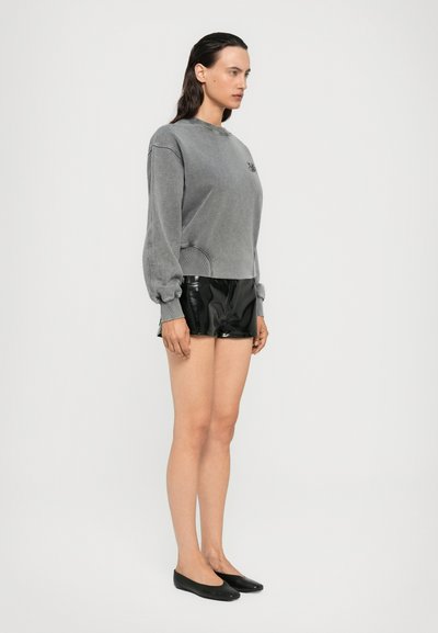 Donna in piedi di lato che indossa una felpa grigia, pantaloni corti neri lucidi e ballerine nere su uno sfondo chiaro e neutro.