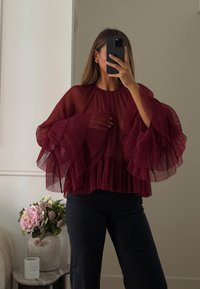 Blusa plissettata borgogna con maniche ampie e voluminose, tessuto texturizzato e collo alto, indossata con jeans scuri su uno sfondo neutro.
