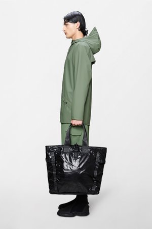 Borsa tote nera lucida con due manici e fibbie, portata da una persona con giacca impermeabile verde e calzature nere, vista di profilo.