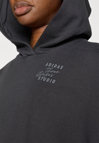 Crna kapuljača sa mekim teksturom. Na prednjoj strani otisnut je logotip "ADIDAS three stripes STUDIO" u svijetlosivoj boji.