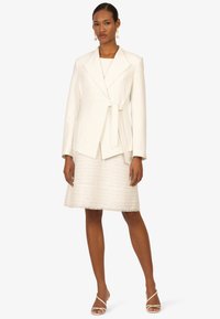 Veste blanche cintrée avec une ceinture à nouer, assortie à une jupe crème texturée avec des détails à franges, et des chaussures minimalistes à lanières.